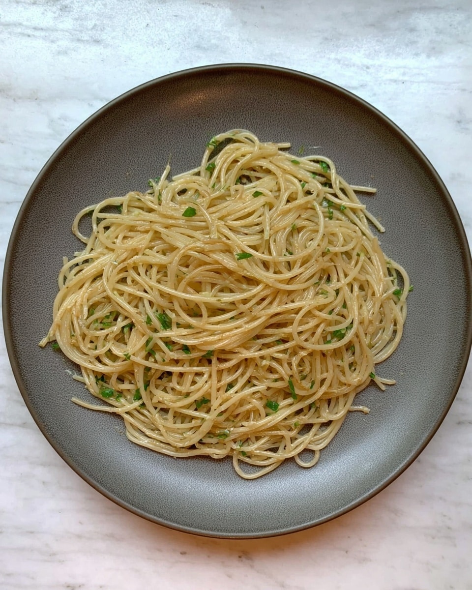 A round white plate holds a single layer of cooked spaghetti noodles. The noodles are light beige with a slight shine from oil or butter and are mixed with small pieces of green herbs scattered evenly throughout. The plate sits on a white marbled textured surface. Photo taken with an iphone --ar 4:5 --v 7