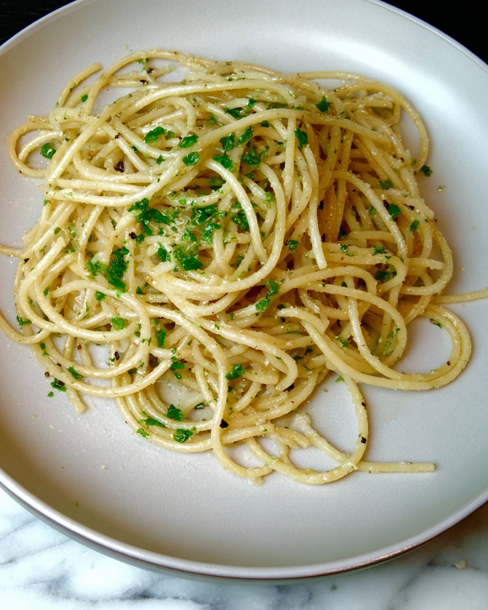 A simple plate of spaghetti pasta is shown, with the noodles loosely piled in the center. The pasta is light yellow with a slight shine, suggesting it is lightly coated with oil or butter. Small green herb pieces are scattered evenly throughout the noodles, giving a touch of color. The spaghetti strands have a smooth texture and are slightly curled and twisted. The food is served on a plain white plate, with a subtle curved edge. The plate sits on a white marbled surface which gives a clean and fresh look. photo taken with an iphone --ar 4:5 --v 7