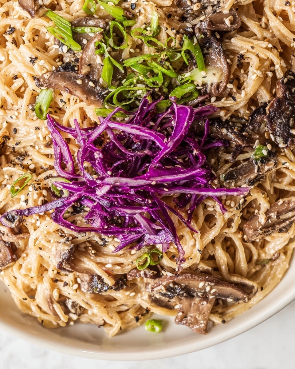 This dish shows a close-up of creamy noodles mixed with slices of brown mushrooms evenly spread through the dish. On top, there are thin strips of bright purple cabbage placed in the upper center, adding a pop of color. To the left of the cabbage, there are chopped green scallions scattered over the noodles. The noodles and toppings are sprinkled with sesame seeds and black pepper, giving texture and specks of white and black all over. The dish is on a white plate on a white marbled surface. Photo taken with an iphone --ar 4:5 --v 7