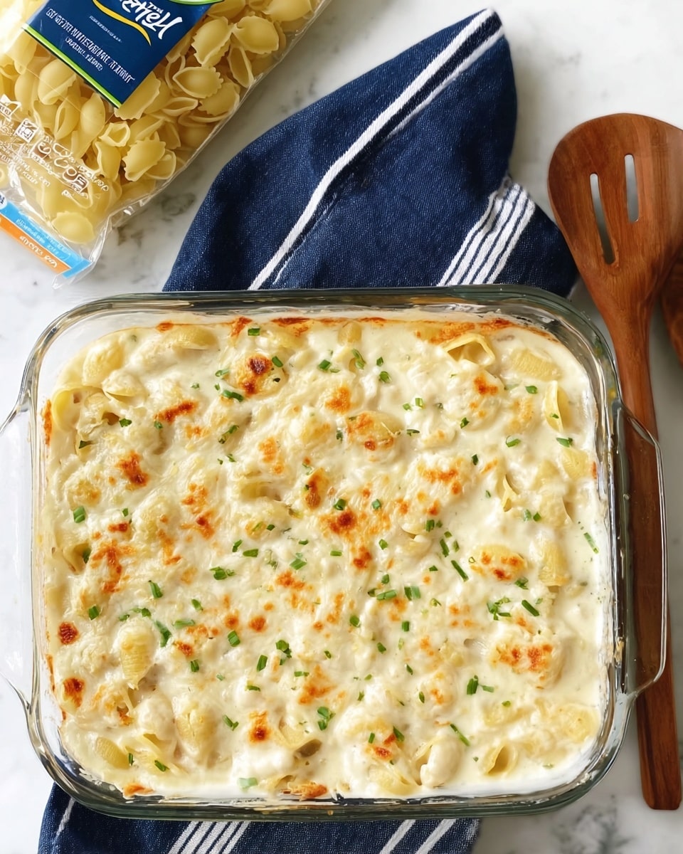 A clear glass baking dish holds a creamy baked pasta casserole with two main layers: the bottom layer is light yellow pasta shells, visible through the top creamy white sauce layer, which has specks of browned spots and is sprinkled with small pieces of green chopped herbs evenly scattered on top. The dish is placed on a white marbled surface with a navy blue cloth with white stripes beside it. There is a wooden slotted spoon on the right, and a partially visible plastic pack of uncooked small shell pasta in the top left corner. photo taken with an iphone --ar 4:5 --v 7