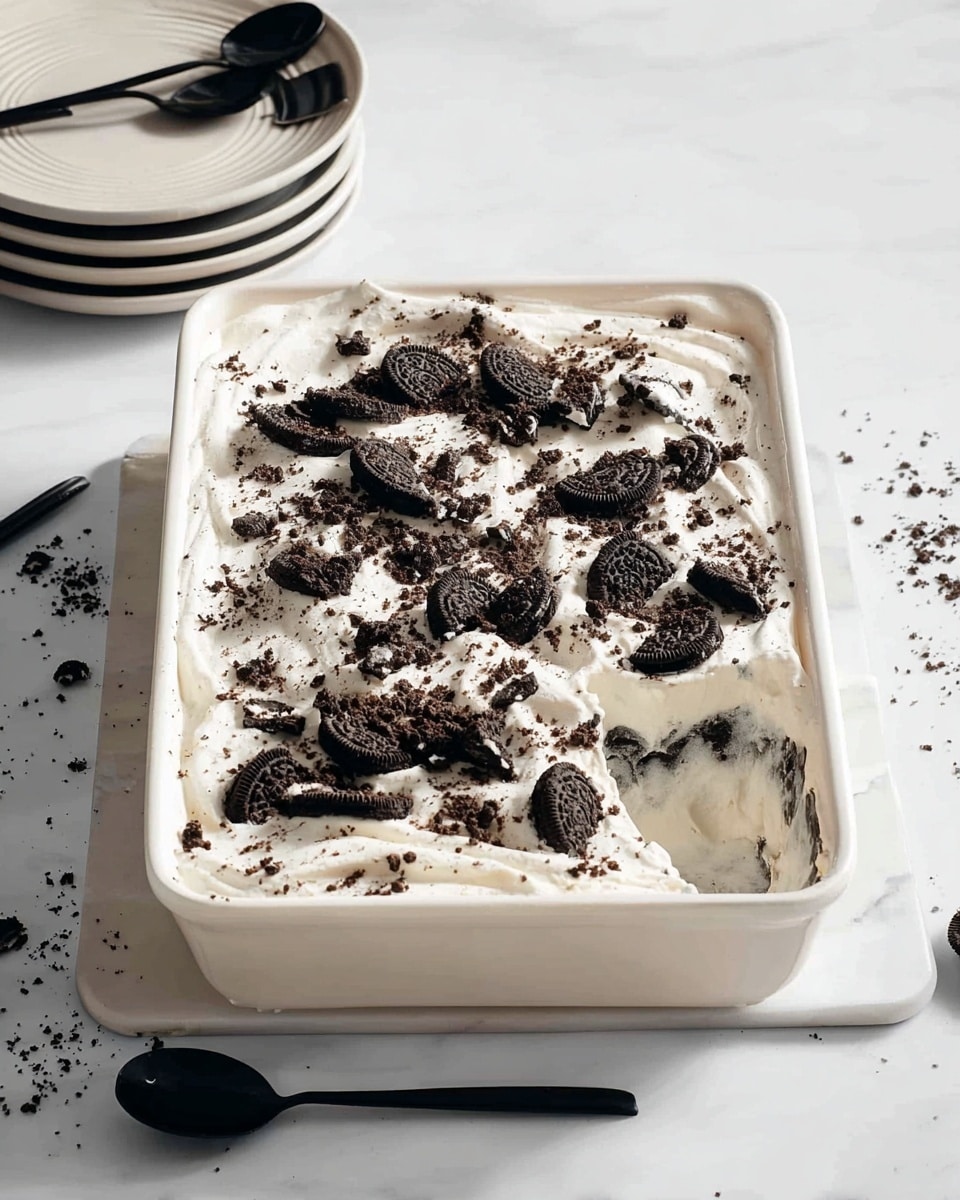 A white rectangular dish filled with a dessert consisting of two clear layers: the bottom layer is hidden beneath a thick, uneven layer of white creamy topping. Crushed black and dark chocolate cookie pieces are scattered generously over the creamy layer, adding texture and contrast. The dish is placed on a white marbled surface with a few crumbs around it, accompanied by a stack of white plates with black spoons on the upper left, and a black spoon placed on the lower right side. Photo taken with an iphone --ar 4:5 --v 7