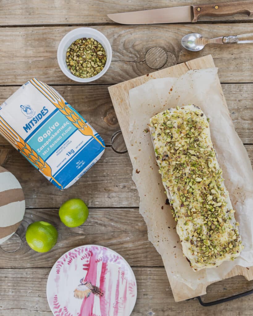 A rectangular loaf cake is placed on a piece of parchment paper on a light wooden board. The cake has a smooth white frosting layer evenly spread on top, covered with a generous amount of chopped green pistachios sprinkled all over, adding texture and color. Near the cake to the top left, there is a small white bowl filled with more chopped pistachios. The background shows a rustic wooden table with visible grains, and other items are scattered around, including a blue and white bag of self-raising flour, two bright green limes, a white plate with pink and red bird and floral designs, and a knife and spoon with a woman's hand holding the spoon at the top right. Photo taken with an iphone --ar 4:5 --v 7