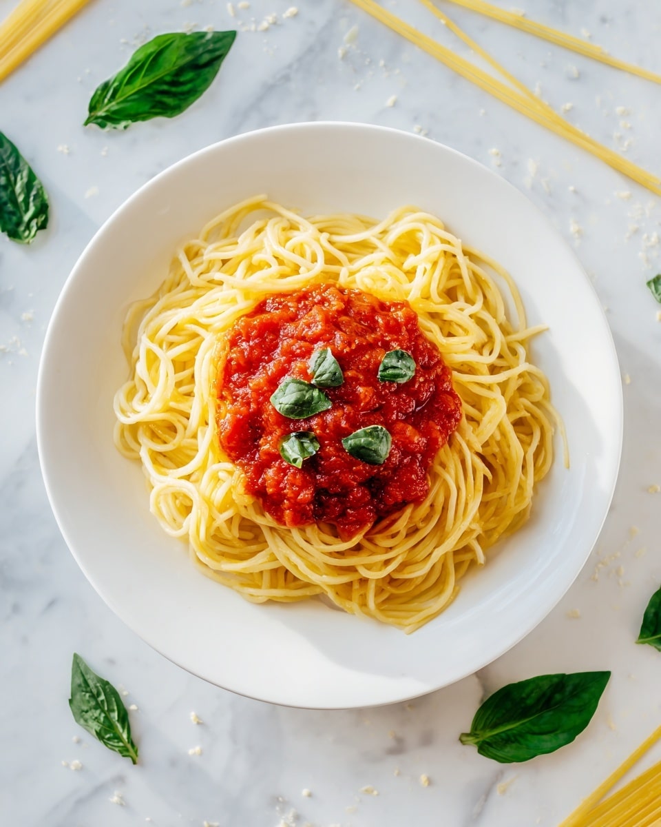 A white plate holds a serving of spaghetti arranged in a loose circular pile, creating a nest-like shape with fine, pale yellow strands. In the center of the spaghetti is a scoop of rich, red tomato sauce, which has a slightly chunky texture. On top of the sauce are small pieces of fresh green basil leaves scattered evenly, adding a touch of color contrast. The plate is set on a white marbled surface, with a few uncooked spaghetti strands and three vibrant green basil leaves placed around the plate as decoration. photo taken with an iphone --ar 4:5 --v 7