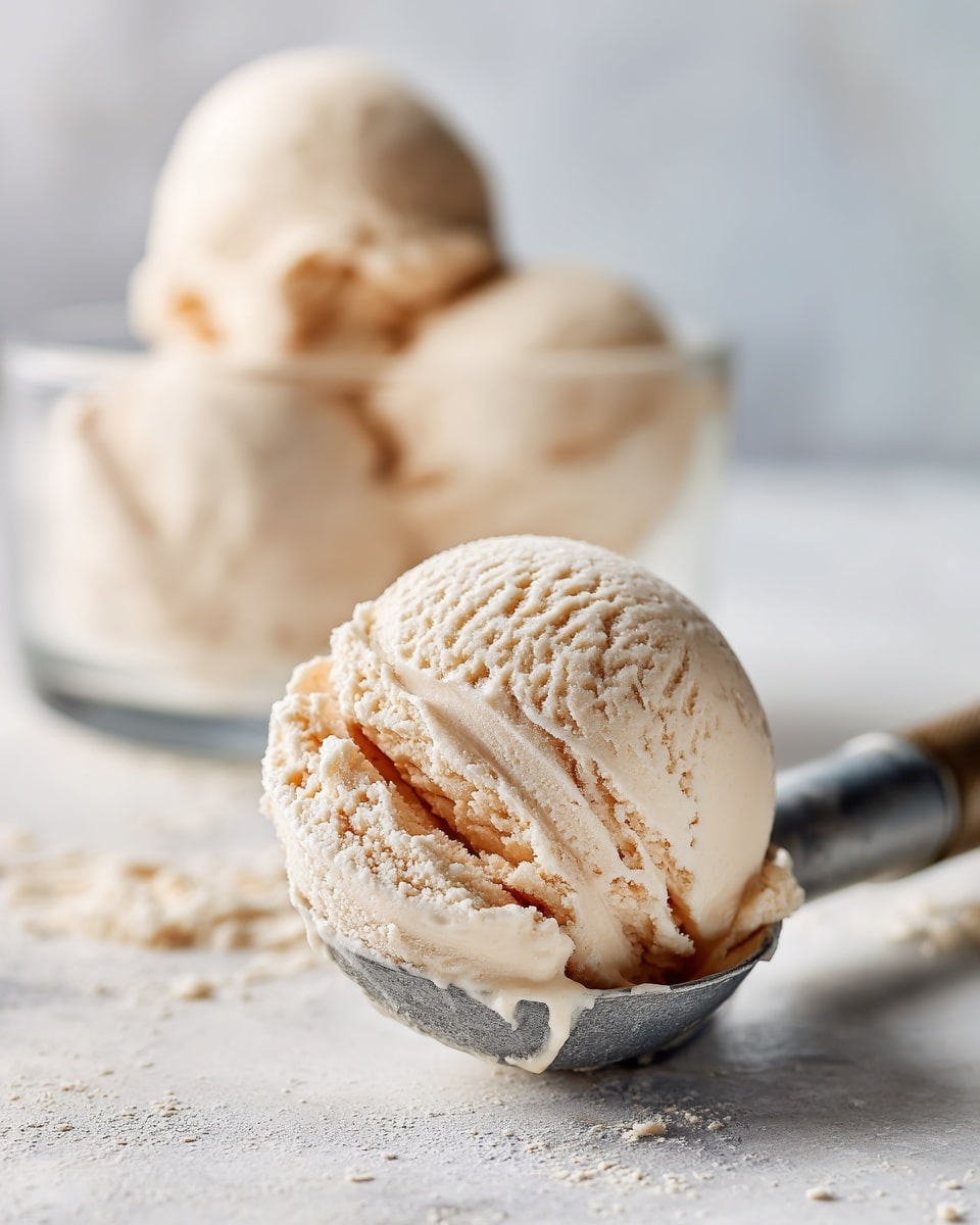 The image shows a single smooth scoop of light beige ice cream sitting on a silver ice cream scooper placed on a white marbled surface. Behind the scoop, there is a clear glass dessert dish filled with three more scoops of the same light beige ice cream, creating a soft and creamy texture look. The background is blurred, highlighting the ice cream texture and color in the foreground. photo taken with an iphone --ar 4:5 --v 7