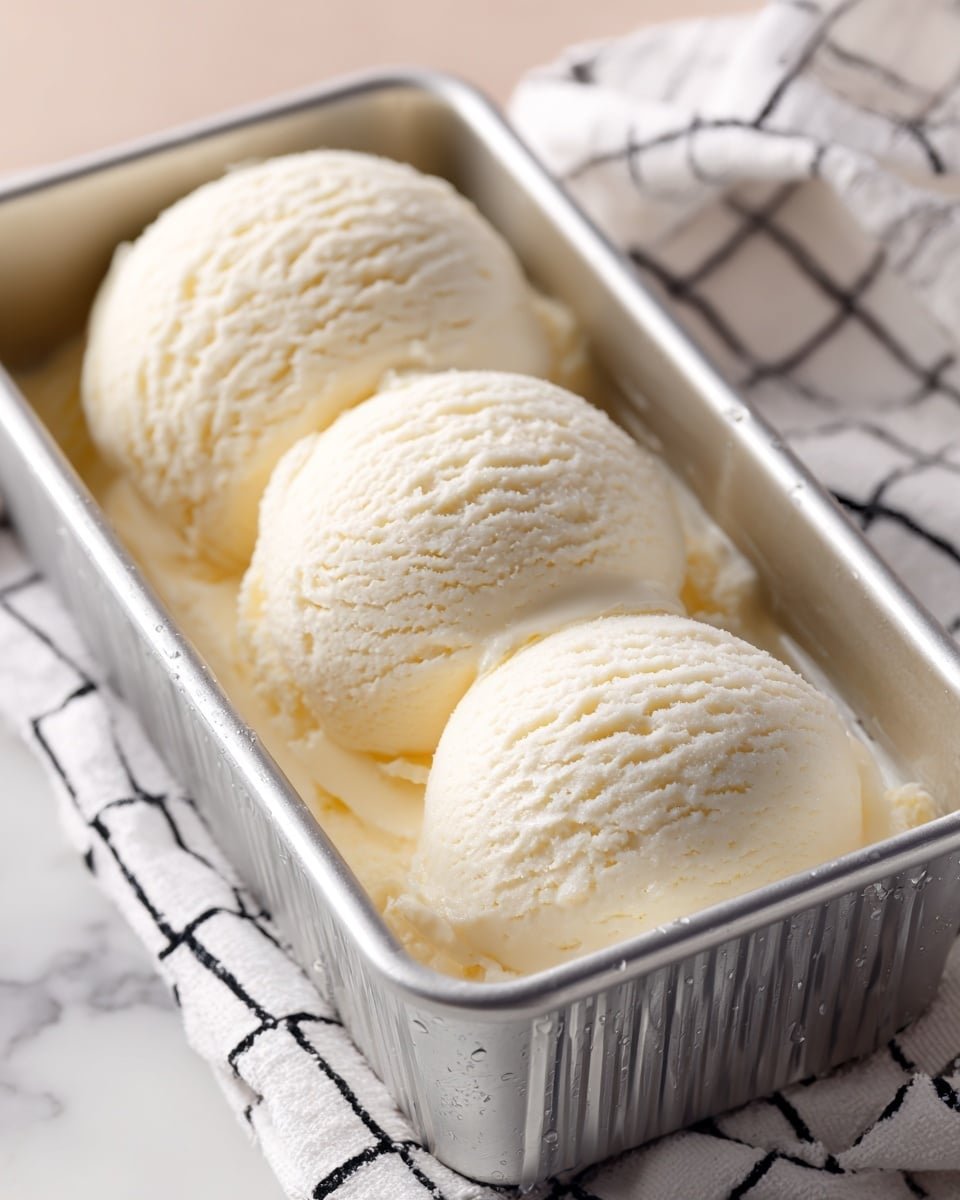Three scoops of light beige ice cream with a creamy texture are stacked inside a metal container. The ice cream's surface looks soft and slightly uneven with some small folds and air pockets visible. The container has a slightly worn look with vertical ridges and is placed on a white marbled surface. Nearby, a few light-colored round biscuits and a white cloth with thin black stripes are partially visible at the bottom right corner of the image. Photo taken with an iphone --ar 4:5 --v 7