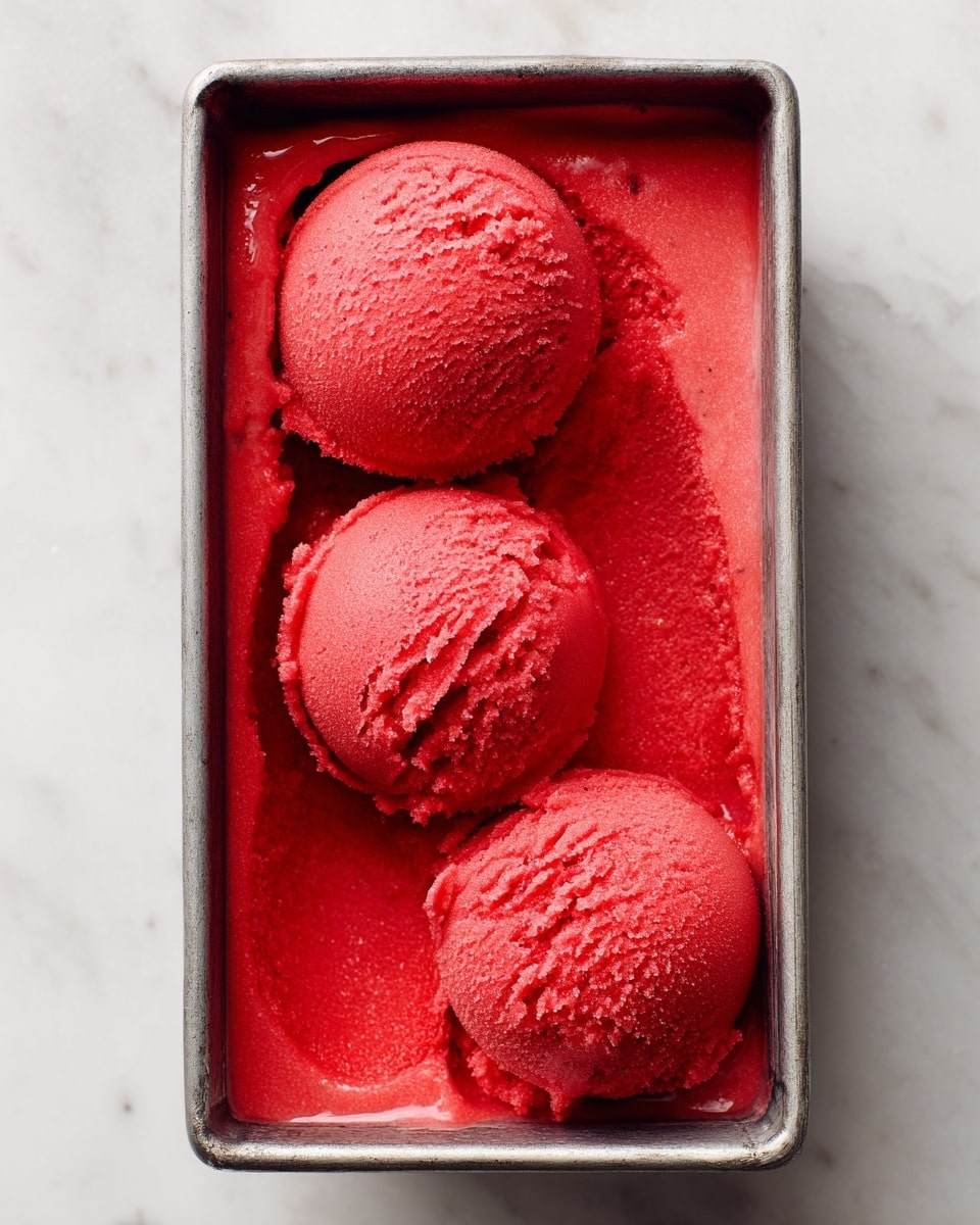 The image shows a metal rectangular container filled with bright red sorbet. Three scoops of sorbet sit on top, each with a smooth but slightly textured surface, showing small cracks and tiny ice crystals. The sorbet inside the container is even and fills the whole space, with some rough edges around the scoops. The background is a white marbled texture. photo taken with an iphone --ar 4:5 --v 7