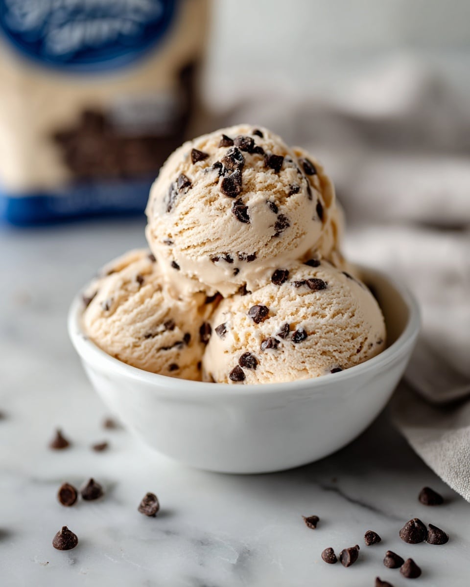 A small white bowl with a gray rim holds three scoops of light brown ice cream mixed with dark brown chocolate chips scattered on top and inside the ice cream. Some extra chocolate chips are placed around the bowl on a white marbled surface. In the background, a blurred white bag with blue and brown details is partially visible. photo taken with an iphone --ar 4:5 --v 7