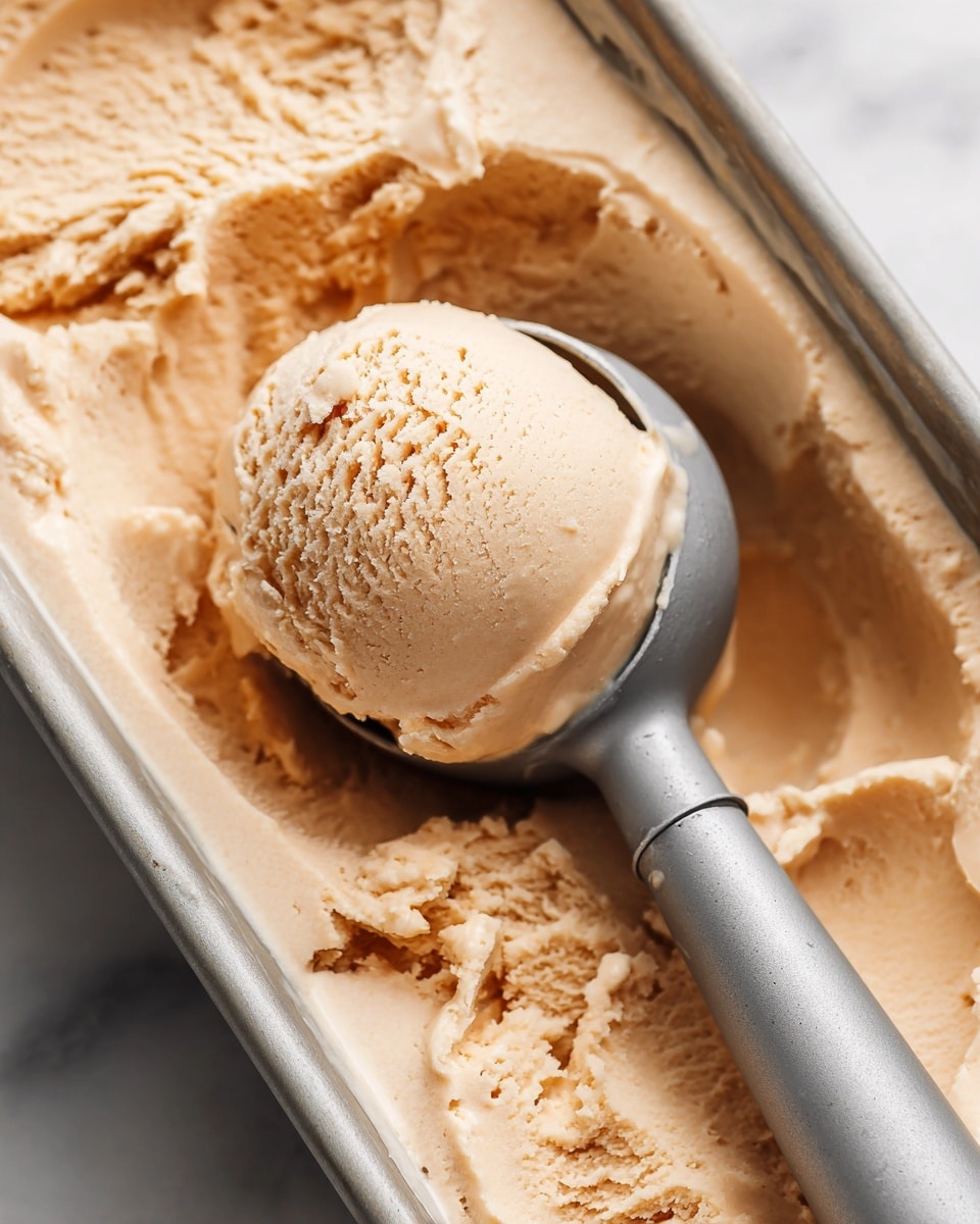A close-up image showing a scoop of light brown ice cream with a creamy, smooth, and slightly soft texture held by a gray ice cream scoop, resting inside a metal container filled with more ice cream of the same color and texture, all placed on a white marbled surface. Photo taken with an iphone --ar 4:5 --v 7