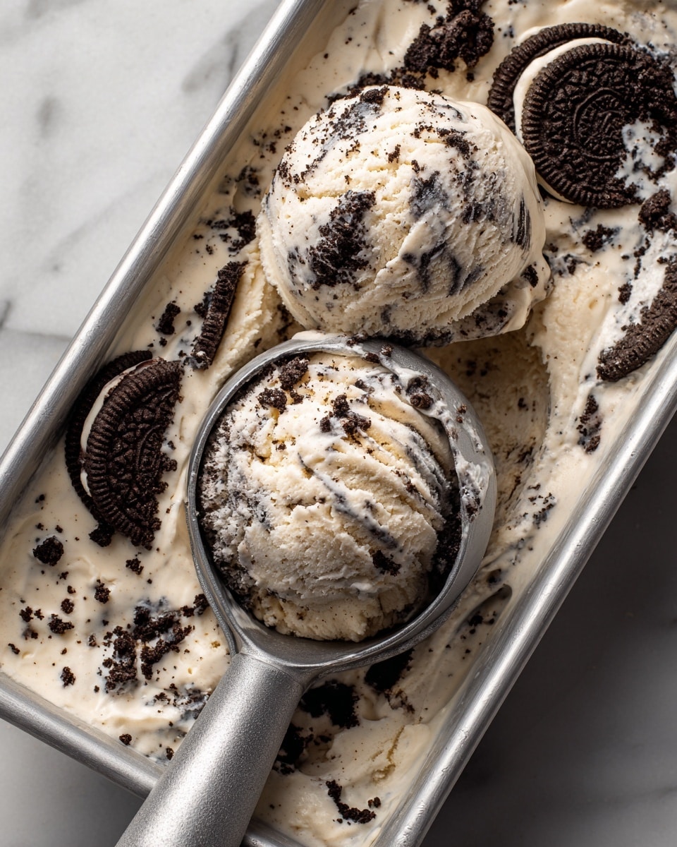 The image shows a close-up of two scoops of cookies and cream ice cream in a rectangular metal container. The ice cream has a creamy white base with many dark brown cookie pieces mixed in, creating a speckled pattern throughout. The scoops are round and smooth with some textural crumbs on top. A gray ice cream scooper is pressing into the ice cream, lifting one scoop partly out of the container. The background surface is a white marbled texture, and the focus is on the rich texture and contrast of the ice cream and cookies. photo taken with an iphone --ar 4:5 --v 7