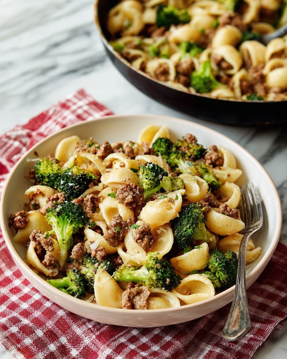 The image shows a close-up of a white bowl filled with three layers: a bottom layer of small, round pasta in a light cream color, a middle layer of cooked small crumbled pieces of brown meat, and a top layer of bright green broccoli florets and leafy greens mixed evenly throughout. The bowl sits on a white marbled surface with a red and white checkered cloth underneath. A large gray pan filled with the same pasta dish is partially visible in the background, along with a fork to the right of the bowl. Photo taken with an iphone --ar 4:5 --v 7