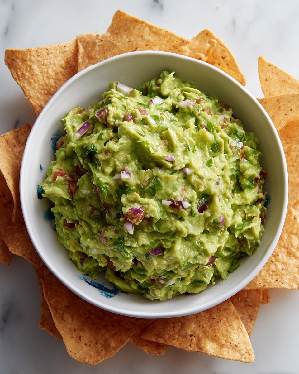 A white bowl filled with chunky guacamole, showing textured layers of mashed avocado mixed with bright red tomato chunks and small pieces of purple onion, all sitting in the center of the bowl. Around the bowl is a ring of golden-yellow tortilla chips, some overlapping and stacked casually. The whole arrangement rests on a white marbled surface, giving a clean and fresh look. photo taken with an iphone --ar 4:5 --v 7