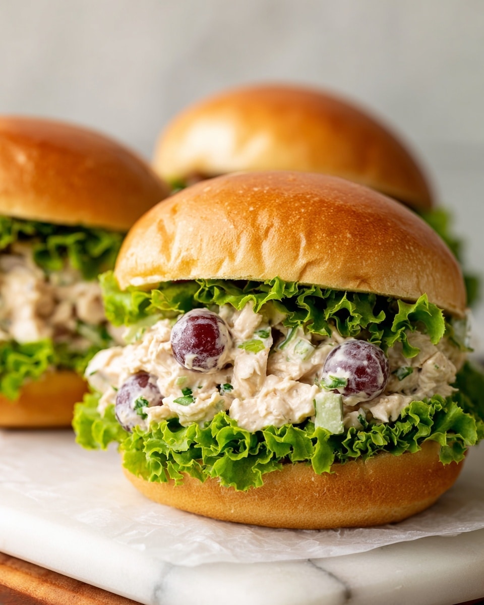 The image shows two stacked sandwiches with soft, shiny brown buns on a white marbled surface. Each sandwich has a layer of bright green leafy lettuce at the bottom, topped with a creamy chicken salad mixed with visible small pieces of light green celery and dark purple grapes. The chicken salad layer is thick, textured, and creamy, filling the buns generously. The buns have a soft, fluffy inside with a light, airy texture. The sandwiches are placed on a piece of white parchment paper on a wooden cutting board. Photo taken with an iphone --ar 4:5 --v 7