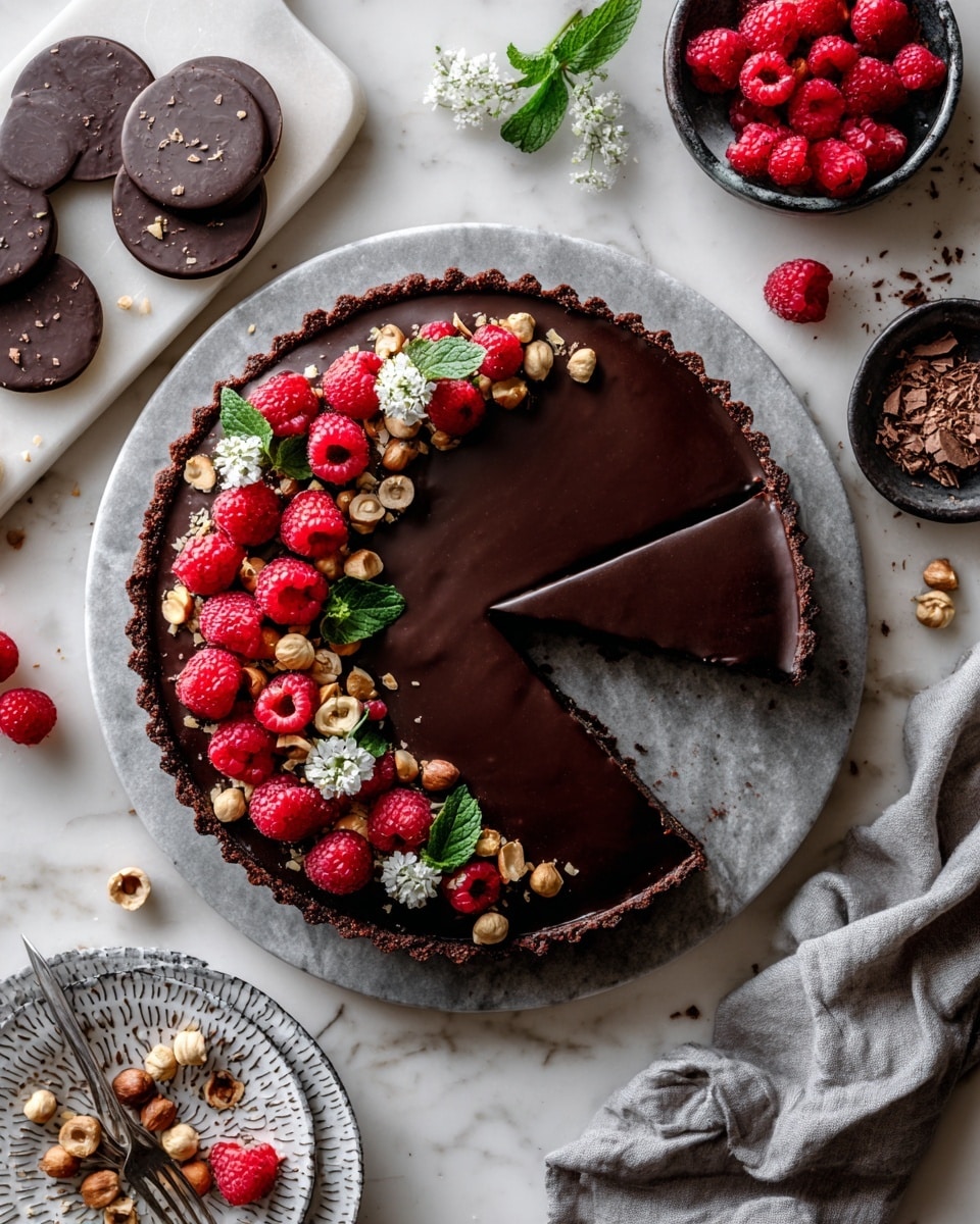 A round chocolate tart with a smooth, dark chocolate filling sits centered on a round grey marble plate. The tart crust is textured dark brown, almost crumbly, and the edge is decorated with a mix of red raspberries, whole hazelnuts, chocolate shavings, and small green mint leaves, covering about half the tart's circumference. Around the tart on a white marbled background are several small bowls and plates holding raspberries, chocolate pieces, hazelnuts, and fresh green mint. A soft gray cloth is casually placed near the tart, adding a cozy touch. Photo taken with an iphone --ar 4:5 --v 7
