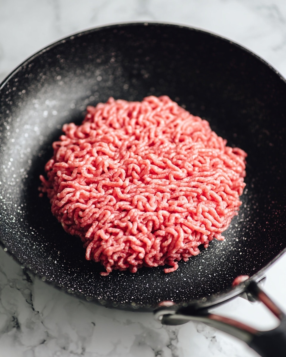 A single layer of raw ground meat is spread flat in a roughly square shape inside a black frying pan with a speckled texture. The meat is bright pink with visible small bits of white fat, showing a stringy pattern from the grinding process. The frying pan sits on a white marbled surface that is slightly blurred in the background. photo taken with an iphone --ar 4:5 --v 7