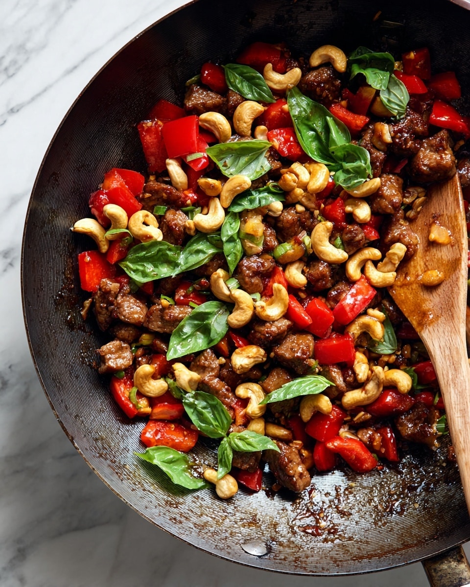 A close-up of a dark cast iron pan filled with a stir-fry dish that has three main layers: the bottom layer is small browned pieces of meat mixed with bright red bell pepper chunks, the middle layer includes golden cashew nuts scattered evenly, and the top layer is fresh green basil leaves placed all over. A wooden spatula is partially visible on the right side inside the pan with a coating of sauce on its surface. The whole image sits on a white marbled background. Photo taken with an iphone --ar 4:5 --v 7