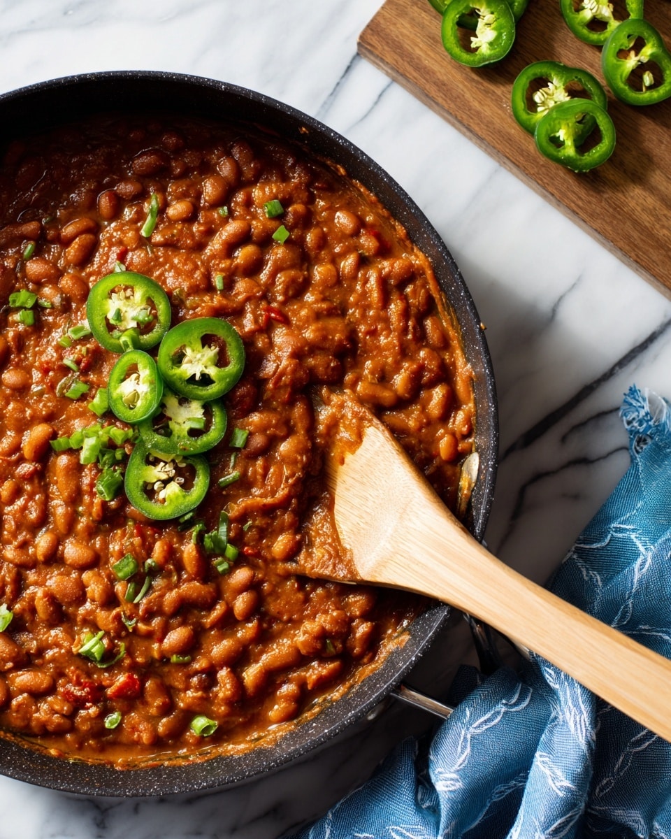 A close-up view of a dark cast iron pan filled with a thick layer of reddish-brown cooked beans in a rich sauce, showing some small chunks of vegetables mixed in. On top, there's a small pile of fresh dark green sliced jalapeño peppers, adding a bright and fresh contrast. To the right, a wooden spoon is partially submerged in the beans, covered in sauce and beans. In the blurred white marbled background, there is a wooden board with two whole green jalapeños and some sliced pieces scattered around, along with a stack of white bowls and a dark blue cloth with a white stitched pattern. Photo taken with an iphone --ar 4:5 --v 7
