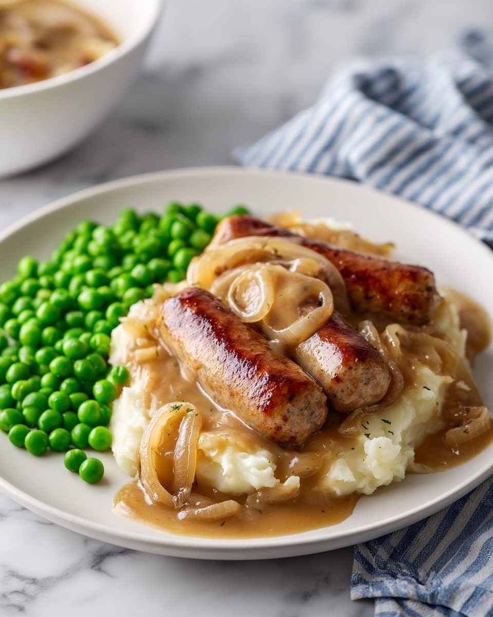 A white plate holds three browned sausages placed on a base of creamy, smooth mashed potatoes that cover most of the plate. The mashed potatoes are topped with a thick brown onion gravy with visible cooked onion slices spread evenly over the sausages and potatoes. On one side of the plate, there is a neat pile of bright green peas, providing a fresh contrast in color. The plate is set on a white marbled surface, with a blurred blue and white striped cloth in the background, and a white bowl is partially visible to the side. Photo taken with an iphone --ar 4:5 --v 7