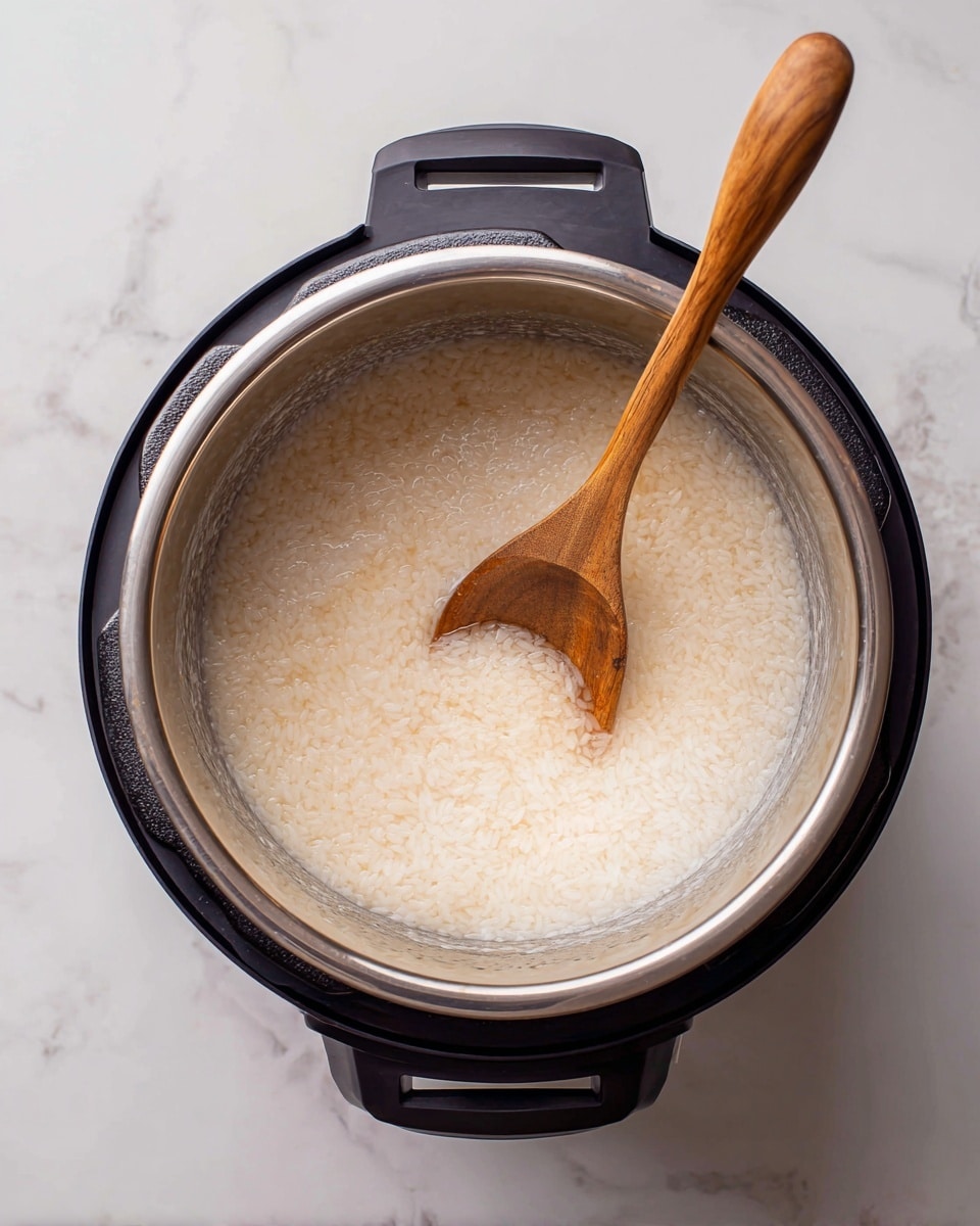 The image shows a top view of a black Instant Pot with a shiny silver inner pot inside. The pot contains a creamy white liquid with grain-like pieces of rice partially submerged and slightly thickened around the edges, blending from light beige to soft cream colors. A wooden spoon with a rounded head lies inside the pot, resting near the center and slightly stirring the rice mixture. The pot is placed on a surface with a white marbled texture. photo taken with an iphone --ar 4:5 --v 7