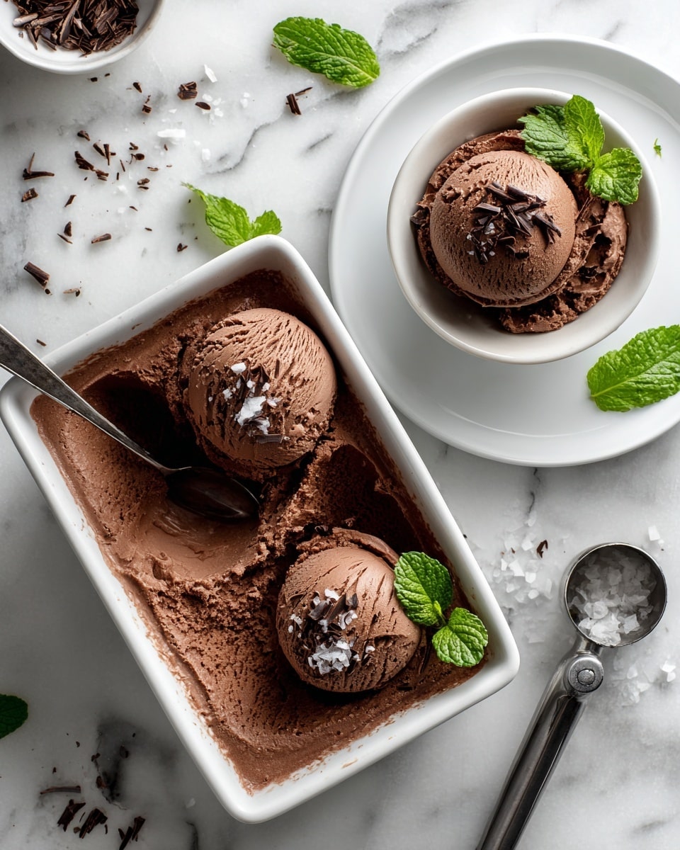 The image shows a rectangular container filled with smooth, rich, brown chocolate ice cream that has a scoop taken from one side, displaying a creamy texture. Above it, two small white bowls with a scoop of chocolate ice cream sit on white plates—one plate with yellow dotted edges and the other plain white. One scoop is topped with small chocolate shavings, while the other has white chocolate flakes on top. Both bowls include a small metal spoon on the side. Scattered mint leaves and tiny chocolate shavings decorate the white marbled surface, along with a metal ice cream scoop positioned on the right side. photo taken with an iphone --ar 4:5 --v 7