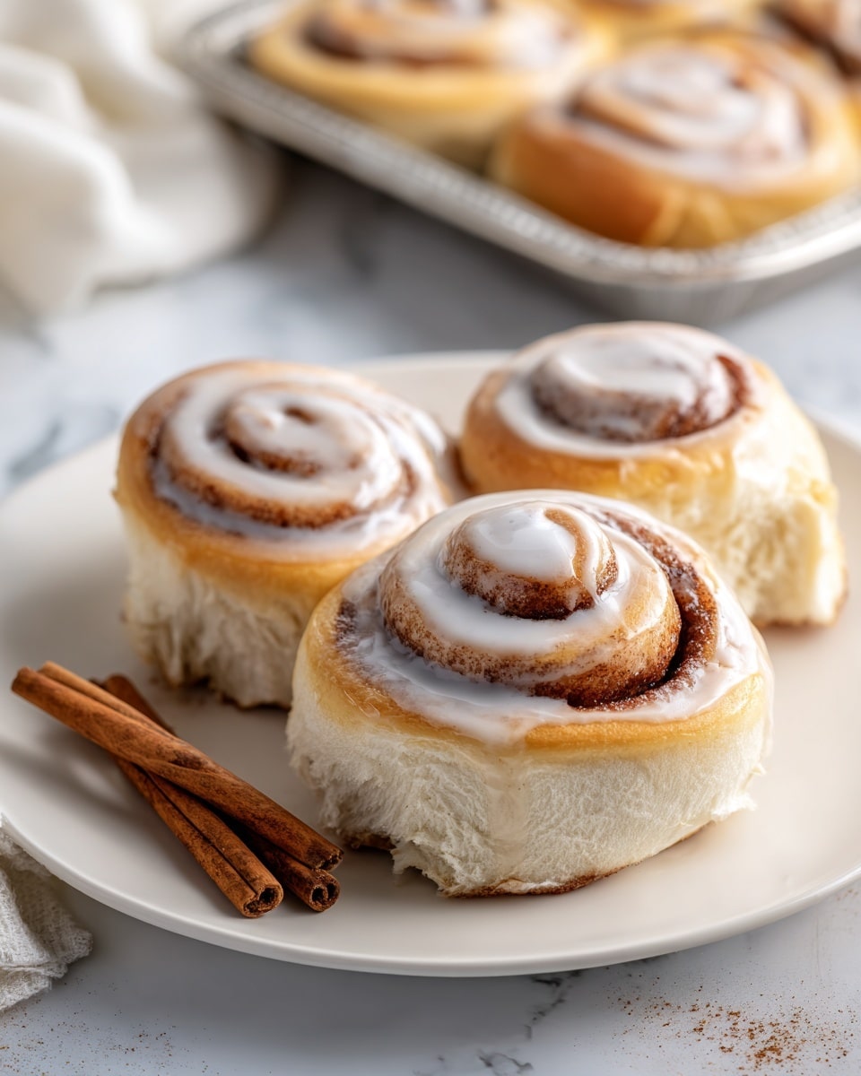 The image shows three cinnamon rolls on a white plate, arranged closely together. Each roll has a light golden-brown dough base with a dark brown cinnamon swirl pattern spiraled through the layers. The rolls have a light, glossy white icing covering the top, clinging to the swirls in patches. Next to the rolls on the left side of the plate, two whole cinnamon sticks lie flat. The plate is set on a white marbled surface with part of a silver tray and a blue cloth with a cinnamon stick blurred in the background. photo taken with an iphone --ar 4:5 --v 7