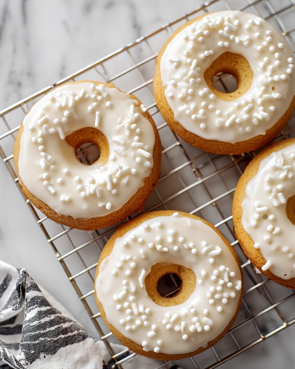 Four donuts with a light golden-brown base are arranged on a silver wire cooling rack. Each donut is topped with a smooth layer of white icing that covers the top surface fully, decorated with small white round and rod-shaped sprinkles scattered evenly. The cooling rack is placed on a white marbled surface with a soft light shining from above, highlighting the texture of the donuts and icing. Part of a white bowl and a striped cloth are visible at the edges of the image. Photo taken with an iphone --ar 4:5 --v 7