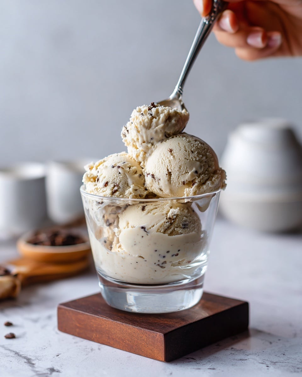 A clear glass dessert cup is filled with three scoops of light brown ice cream that shows small dark specks, giving it a textured look. The scoops are creamy and slightly rounded, with one scoop being lifted by a silver spoon held by a woman's hand from the top right. The dessert cup is placed on a small dark wooden block, and the background features a white marbled surface with soft blurred white dishes and fabric scattered around. photo taken with an iphone --ar 4:5 --v 7