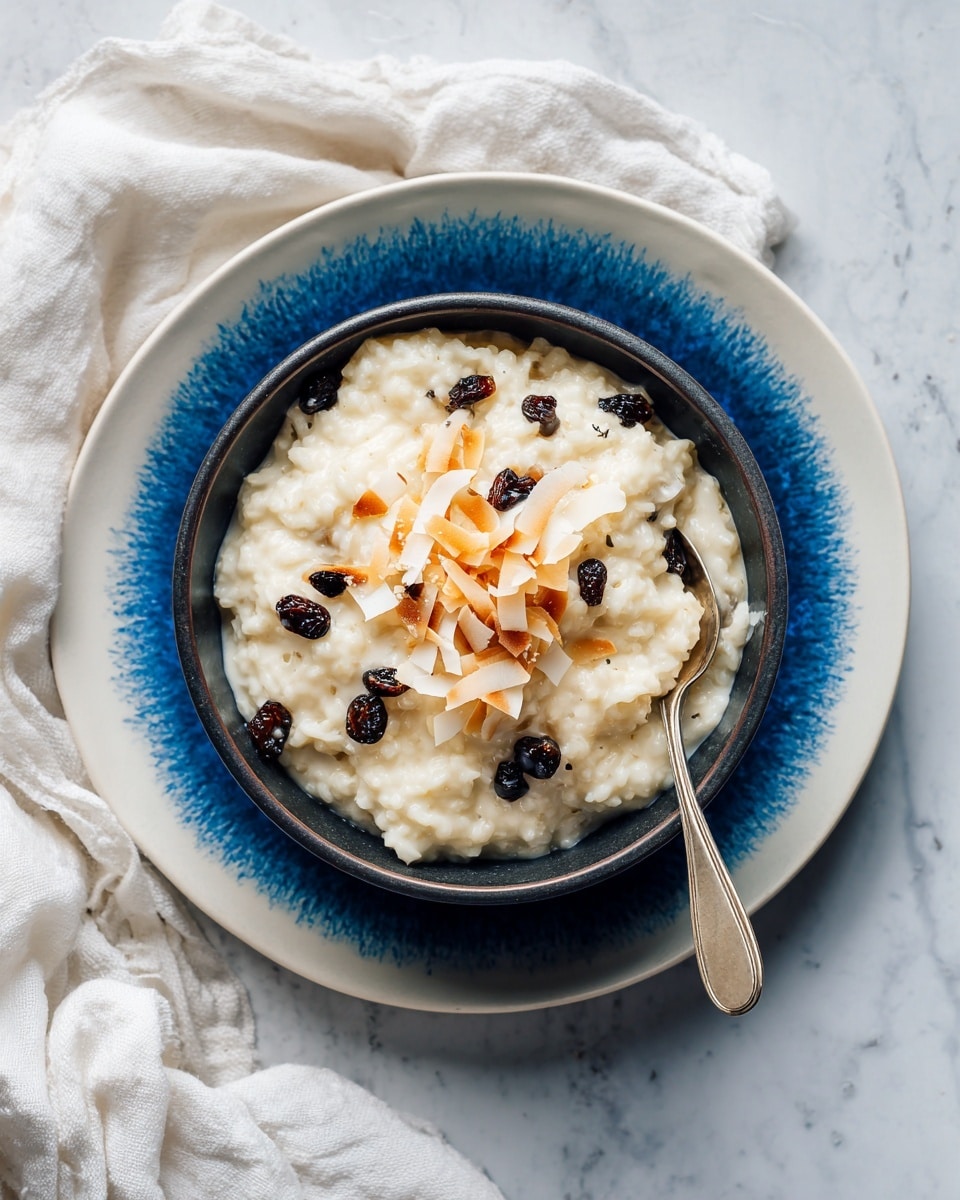 A blue bowl holds a smaller gray bowl filled with creamy rice pudding mixed with dark raisins and topped with scattered toasted coconut flakes. A silver spoon rests inside the smaller bowl on the right side, partially immersed in the pudding. The bowls are placed on a white marbled surface with a soft white cloth partially visible in the upper left corner. The lighting is natural and soft, highlighting the textures of the pudding and toasted coconut flakes photo taken with an iphone --ar 4:5 --v 7