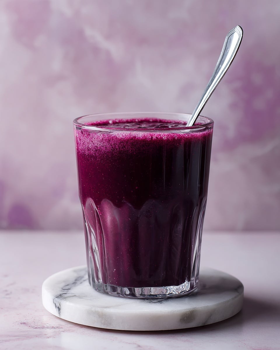 A clear glass cup filled with a thick, smooth bright purple smoothie, nearly full to the top. A metal spoon with a dotted handle is placed inside the cup, slightly dipping into the smoothie, causing soft ripples and reflections on the surface. The cup sits on a small, round white marbled coaster, all placed on a white marbled surface. In the blurred background, hints of pink and purple elements are visible, adding a soft and cozy feel to the image. Photo taken with an iphone --ar 4:5 --v 7