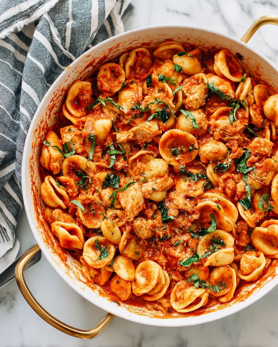 A white pan filled with one layer of small round pasta shells coated in a rich orange-red tomato sauce. Mixed in are pieces of light golden cooked chicken and small chunks of red tomatoes. The pasta is topped with scattered bright green fresh basil leaves, adding color contrast. The pan has two gold handles, and the background is a white marbled texture. Photo taken with an iphone --ar 4:5 --v 7