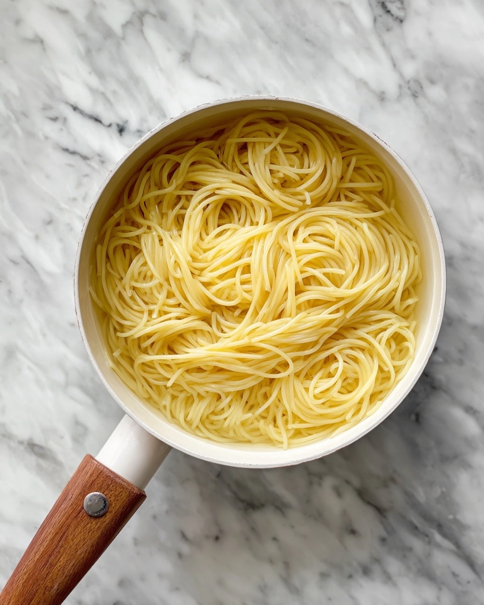 The image shows a white pot filled with cooked long spaghetti noodles sitting in clear water. The spaghetti is light yellow and tangled neatly inside the pot. The pot has a wooden handle on the left side, resting on a surface with a white marbled texture. The background is a similar white marbled surface with some subtle dark veins. photo taken with an iphone --ar 4:5 --v 7