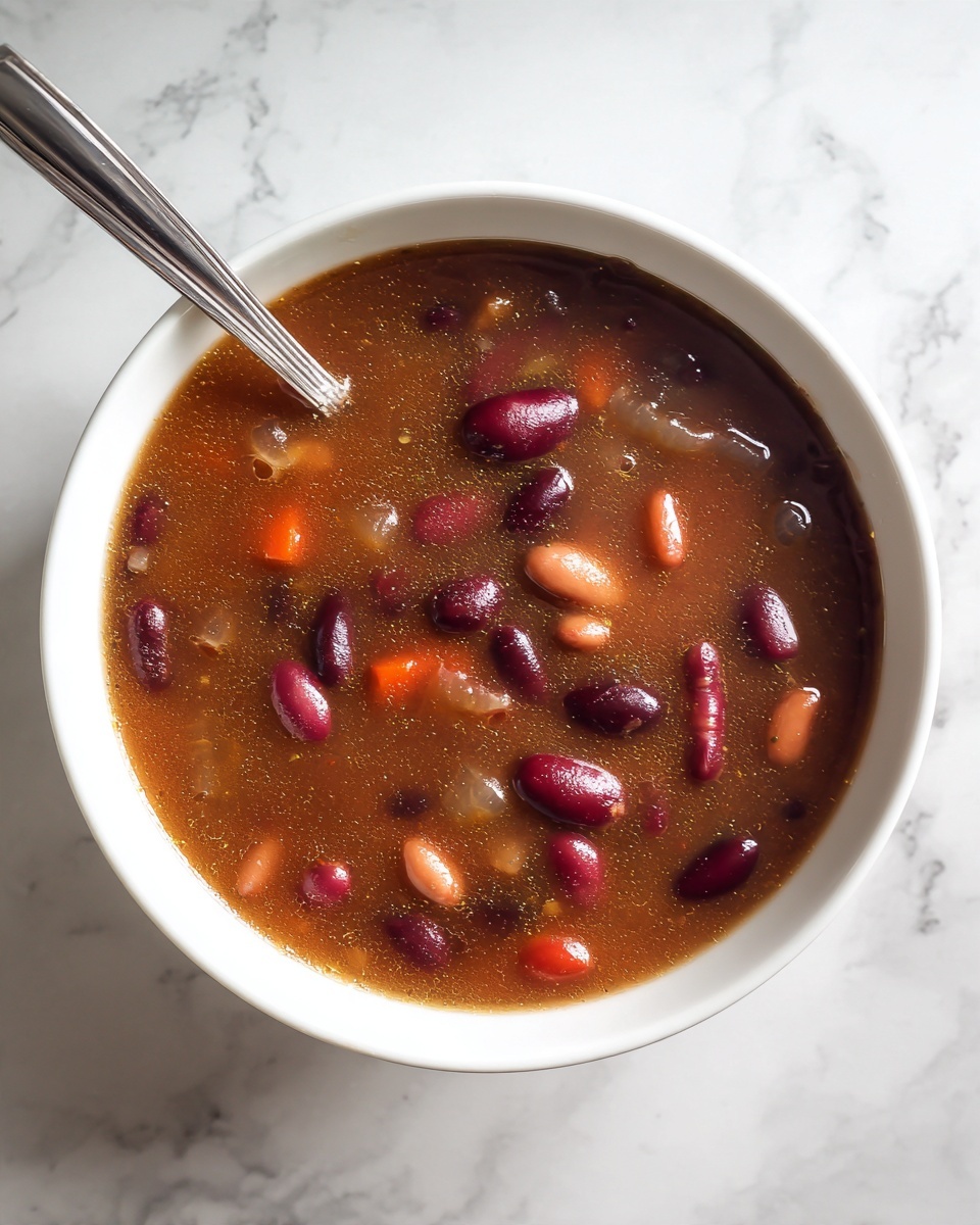 New England Baked Beans Recipe 5 A white bowl filled with dark brown bean soup with a glossy surface. The soup is thick with many whole kidney beans floating on top, showing red, purple, and orange colors, giving a rich texture. A metal spoon rests inside the bowl, placed on the right edge. The bowl is on a white marbled surface, creating a clean and bright background. photo taken with an iphone --ar 4:5 --v 7