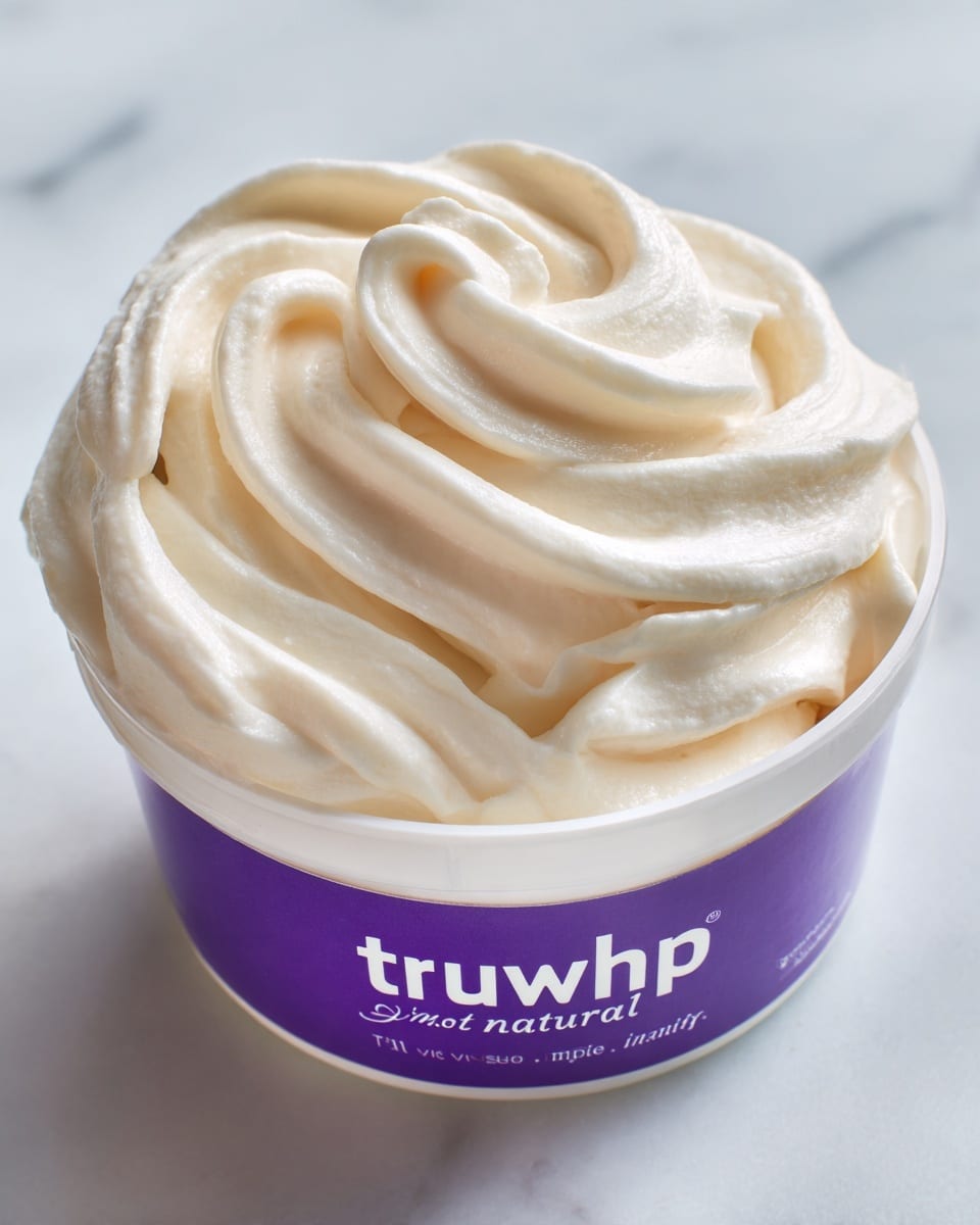 A close-up of an open white container filled with smooth, creamy off-white whipped topping. The whipped topping has soft swirls and peaks on the surface, showing a light and airy texture. The container has a purple label with white text that reads