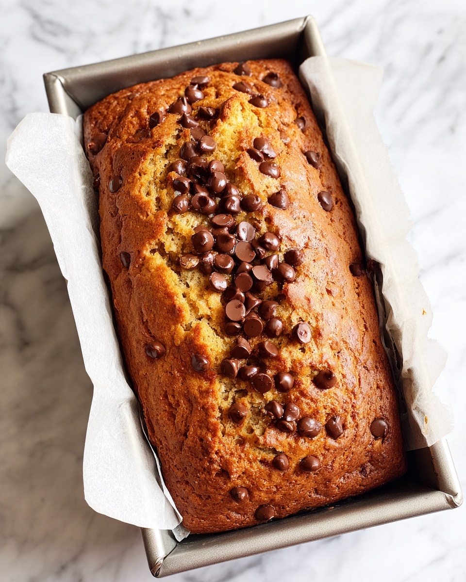 A rectangular baked banana bread loaf in a metal pan lined with parchment paper, showing a light brown, slightly rough textured top with many dark brown chocolate chips spread mainly in a thick line across the middle. The edges of the bread look golden and firm, while the pan rests on a white marbled surface. photo taken with an iphone --ar 4:5 --v 7