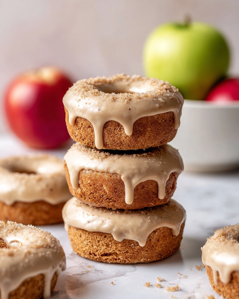A stack of three light brown donuts with a smooth, creamy light beige glaze that drips slightly down the sides, sprinkled with a fine golden powder, is placed on a white marbled surface. Surrounding the stack are additional glazed donuts of similar size and color, arranged casually in the background. A white bowl with a scalloped edge holds bright red and green apples, blurred softly in the background. The overall scene is bright and inviting. photo taken with an iphone --ar 4:5 --v 7