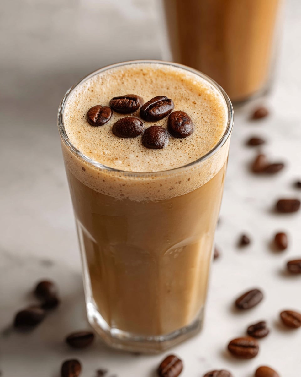 A tall clear glass is filled with a smooth, creamy brown coffee drink topped with seven dark brown coffee beans resting on the surface. The drink has a light frothy texture with small bubbles around the edges. Coffee beans are scattered on a white marbled surface around the glass, and a second similar glass is blurred in the background. photo taken with an iphone --ar 4:5 --v 7