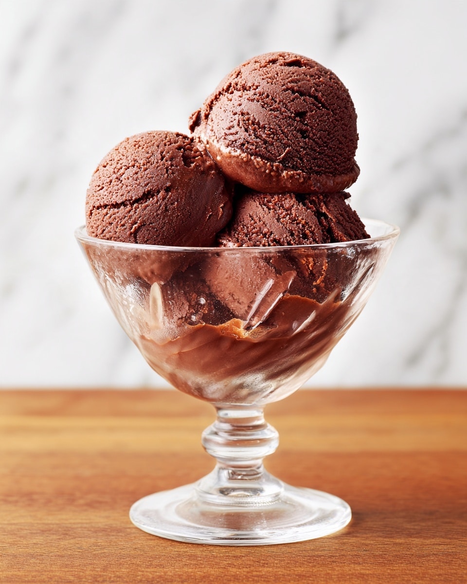 The image shows three scoops of chocolate ice cream stacked in a clear glass dessert dish with a wide, flared top and a sturdy base. The ice cream has a smooth but slightly textured surface and a rich brown color. The glass dish sits on a wooden surface, though the background has been changed to a white marbled texture. The dessert is centered and fills the frame. Photo taken with an iphone --ar 4:5 --v 7